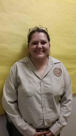 Custodian in brown custodial uniform smiling. She is in from of a yellow backdrop. 